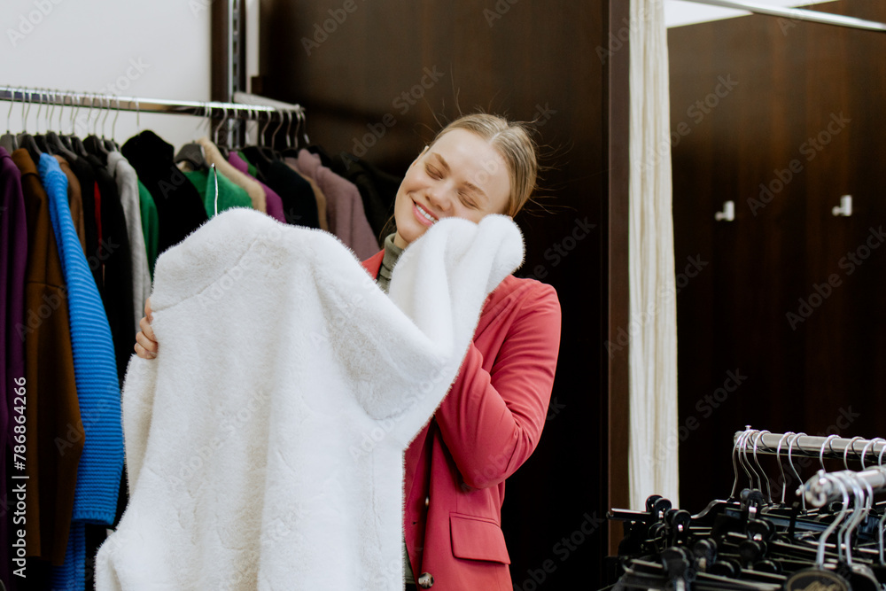 Happy woman touches a white faux fur coat and tries it on for herself, in a women's clothing store, in a boutique. Shopping in a clothing store. Happy woman touches a white faux fur coat and tries it on for herself, in a women's clothing store, in a boutique. Shopping in a clothing store.