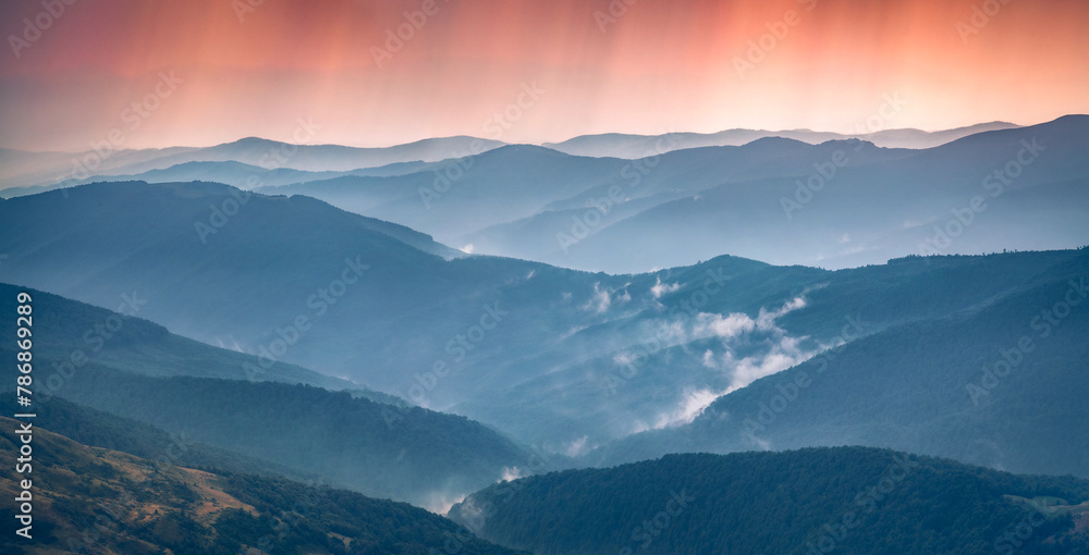 Obraz premium Rainy summer scenery. Silhouettes of rolling hills are covered with deciduous forest. Dramatic summer view of dark blue mountains at sunset. Beauty of nature concept background.