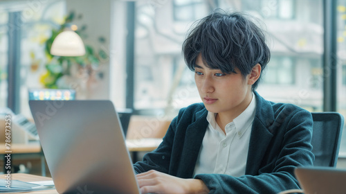 A young asian man works on a computer while sitting at a table in a modern bright office Free Download A young asian man works on a computer while sitting at a table in a modern bright office - GFXLoad