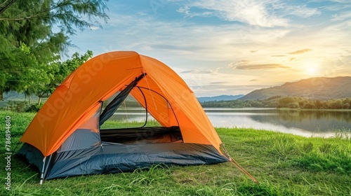 An orange tent is pitched on the green grass next to a serene lake on a sunny day
