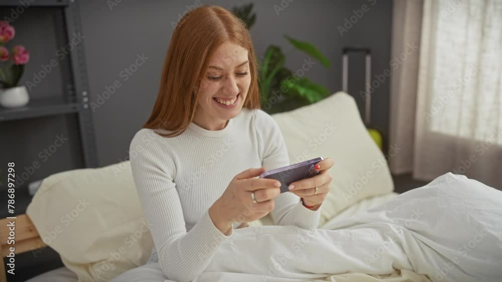 Redheaded woman smiling while texting on her phone in a cozy bedroom setting Redheaded woman smiling while texting on her phone in a cozy bedroom setting