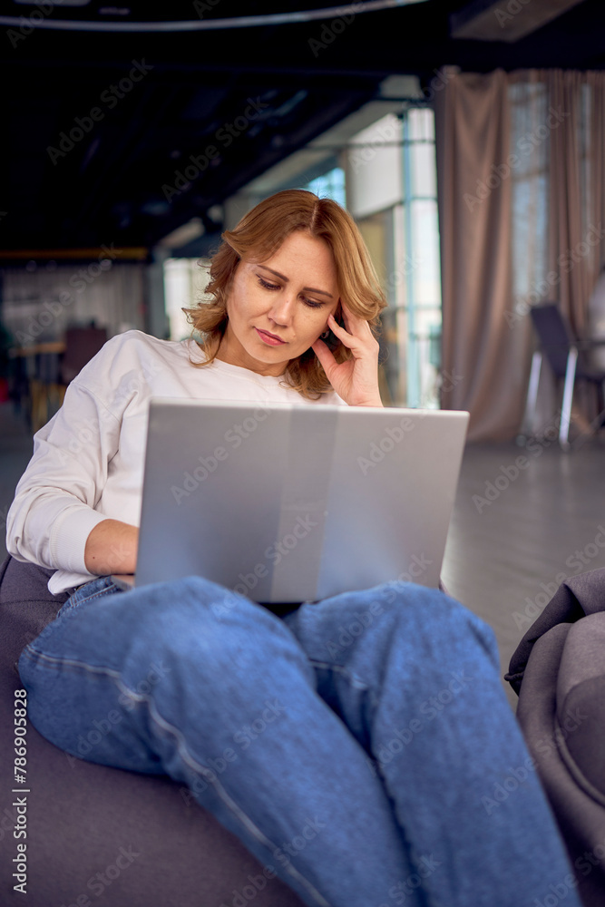 Woman on a puff chair working on her laptop in coworking with modern interior design, neutral tones and green plant Woman on a puff chair working on her laptop in coworking with modern interior design, neutral tones and green plant