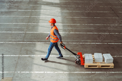 Factory worker, engineer wearing hard hat, vest, work clothes carrying pallet truck with boxes