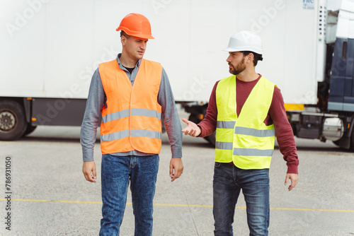 Serious men wearing hard hats and vests talking to each other on the street, near cargo terminal
