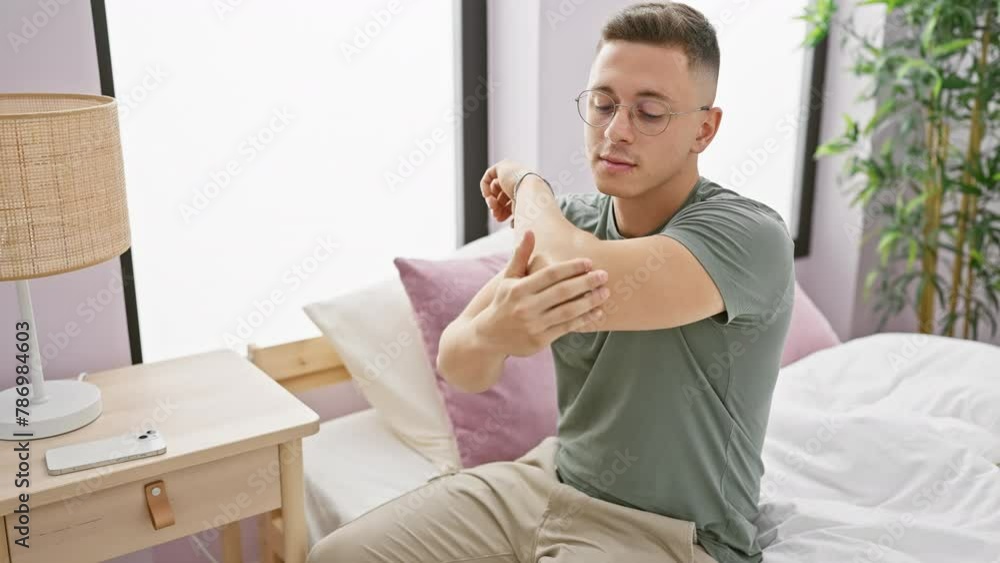 A young hispanic man applies cream to his arms while sitting on his bed in a well-lit bedroom A young hispanic man applies cream to his arms while sitting on his bed in a well-lit bedroom