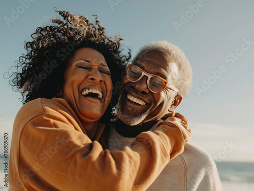A senior africanamerican couple hugging each other on the beach, smiling and wearing sweatshirts with clear blue sky in background. The woman has curly hair and is laughing as she hugs her husband who photo