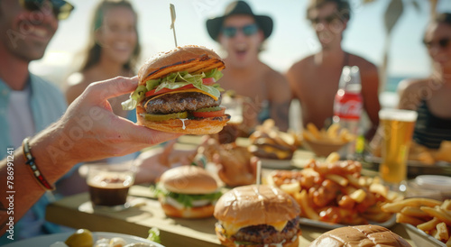 A group of friends enjoying delicious food and drinks at the beach  laughing together while sitting on sun loungers near an outdoor table with burgers and fries.