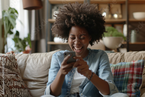 young Afro-American woman sits on a sofa at home, holding a smartphone device, her excitement palpable as she gazes at the mobile screen and gestures yes