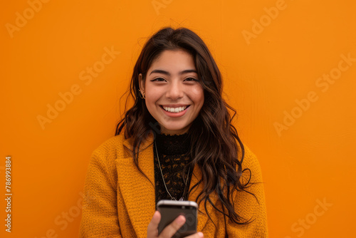 An enthusiastic Latina woman, in her late 20s, smiles warmly as she recommends a smartphone app on a vibrant orange background, demonstrating her tech expertise and engaging communication style.