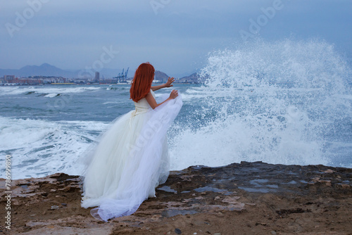 Mujer pelirroja con vestido de novia en la playa y salpicaduras de ola de mar con gesto de parar el splash, Alicante, España