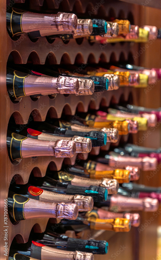 Champagne or cava bottles of different metallic colored labels lined up and stacked on wooden shelves of a wine rack in a gourmet liquor store from a luxury private collection.