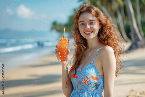 Cheerful smiling woman with long curly hair in blue dress holding an orange summer cocktail on a tropical beach. Happy beach vacation.