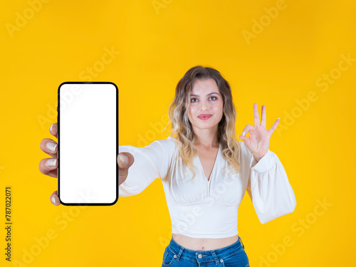 Mobile offer concept, beautiful caucasian blonde curly hair young woman holding showing empty blank screen mobile phone mock up close up to camera. Showing OK sign gesture. Yellow background.