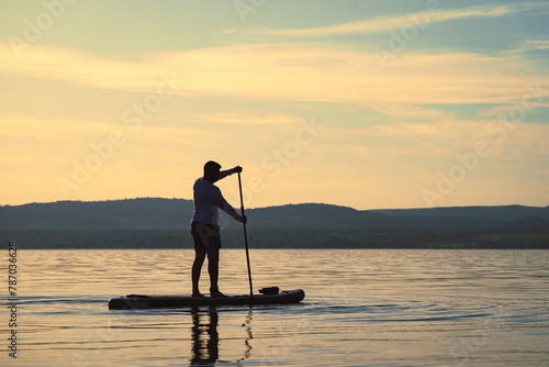 Silhouette of a man paddling on a stand up paddle boarder or SUP on the lake at sunset. Hills in the background. Active lifestyle. © Skatty