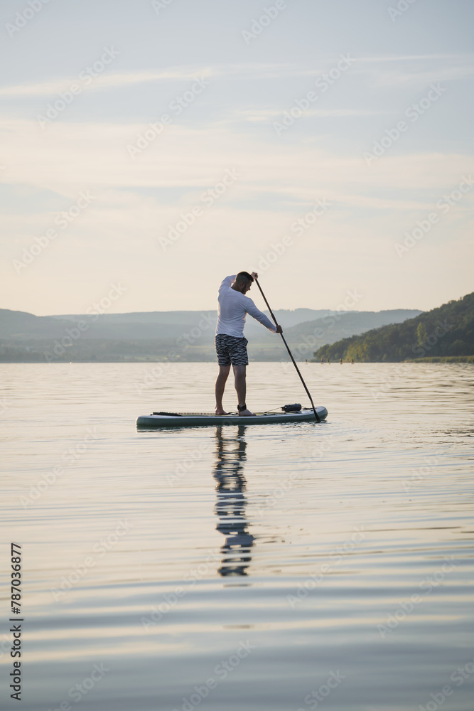 A man is training SUP board on the lake. Stand up paddle boarding. Hills in the background. Enjoying the vacation. Active lifestyle.