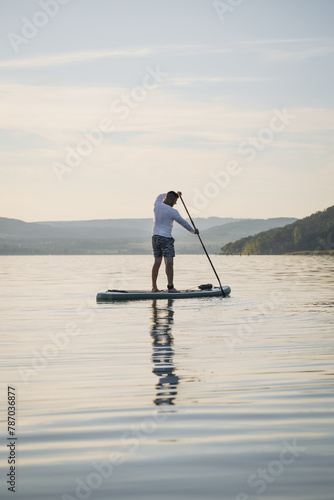 A man is training SUP board on the lake. Stand up paddle boarding. Hills in the background. Enjoying the vacation. Active lifestyle. © Skatty