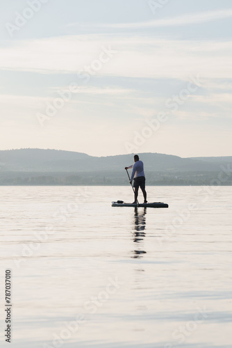 A man is standing and paddling on a paddleboard on the water. Enjoying the vacation. Hills in the background.