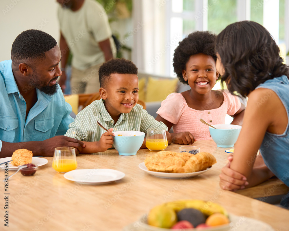 Family Indoors At Home At Kitchen Counter Eating Breakfast With Grandparents In Background Family Indoors At Home At Kitchen Counter Eating Breakfast With Grandparents In Background