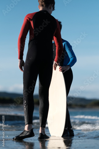 Junge Brüder im Neoprenanzug mit dem Skimboard am Strand im Meerwasser auf Sardinien mit Wellen im Hintergrund