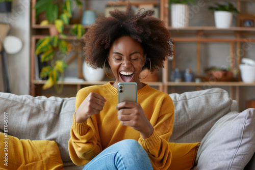 With excitement, a happy black woman sits on a sofa at home, holding a smartphone device, her satisfied expression evident as she gazes at the mobile screen and gestures yes with a clenched fist photo