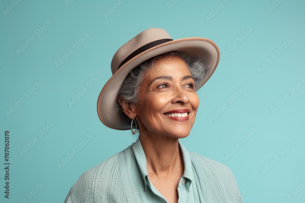 Portrait of a smiling indian woman in her 60s donning a classic fedora while standing against pastel teal background Portrait of a smiling indian woman in her 60s donning a classic fedora while standing against pastel teal background