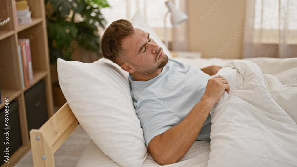 A bearded young man in a blue shirt looks uncomfortable in bed, indicating sickness or insomnia in a cozy bedroom setting. A bearded young man in a blue shirt looks uncomfortable in bed, indicating sickness or insomnia in a cozy bedroom setting.