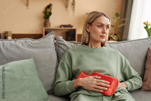 Portrait of adult woman holding red hot water bottle to stomach suffering from period cramps sitting on couch at home copy space