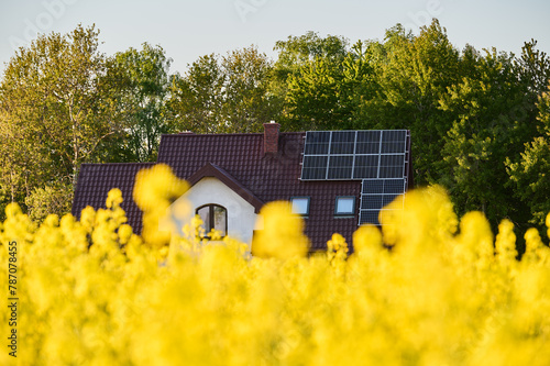 Photovoltaic panels for the production of electricity on the roof of a single-family house next to a field of yellow-flowering rapeseed