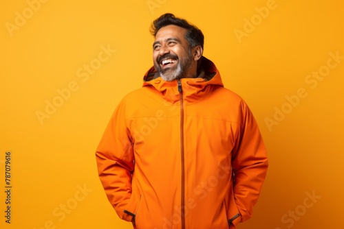 Portrait of a joyful indian man in his 40s wearing a windproof softshell while standing against soft orange background