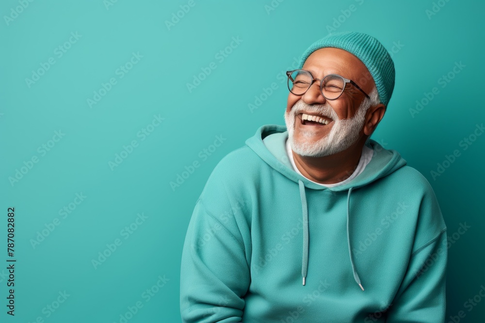 Obraz premium Portrait of a smiling indian man in his 80s sporting a comfortable hoodie in front of soft teal background