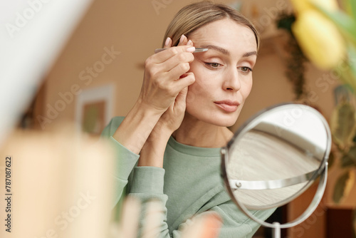 Warm toned portrait of elegant adult woman plucking eyebrows with tweezers looking in mirror at home copy space