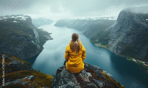 Portrait of a woman in a yellow raincoat sitting from her back on a troll's tongue high in the mountains of Norway against the background of fjords photo