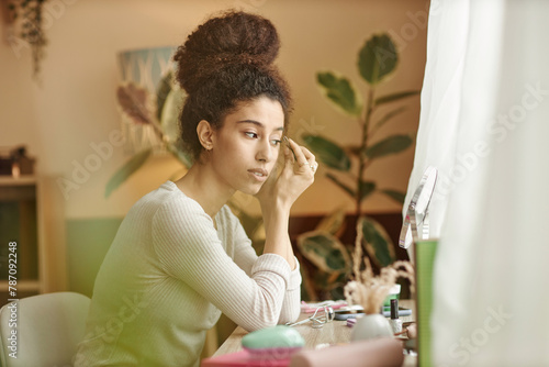 Side view portrait of young Black woman shaping eyebrows with tweezers looking in mirror at home copy space