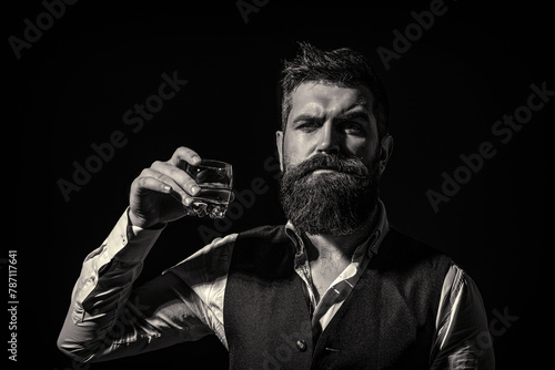 Man with beard holds glass with alcohol. Barman or degustator with curious face holds glass of cognac. Bearded man wearing suit and drinking whiskey, brandy, cognac. Black and white