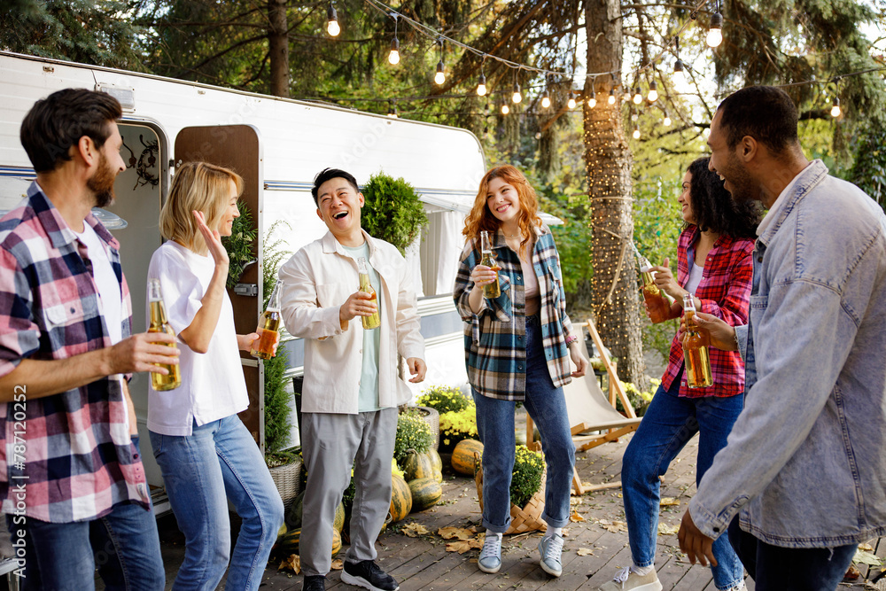 A cheerful group of friends rest in nature near a tourist trailer. Mixed-gender and multinational company of friends toasting, dancing, drinking drinks, have fun.