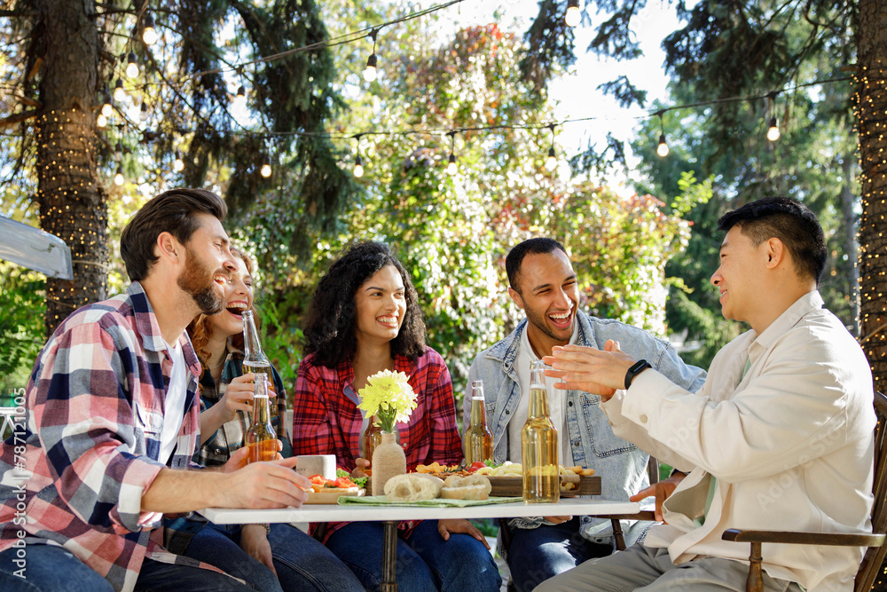 Obraz premium A cheerful multinational company is having fun on a picnic. Multisex friends sitting at the table outdoors celebrating eating and drinking drinks.