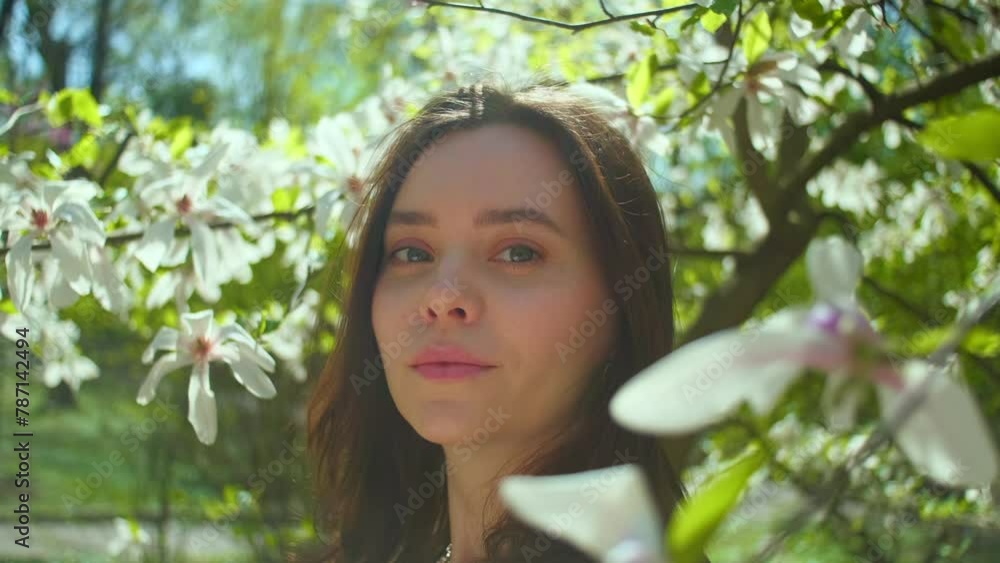 Beautiful woman smiling looks at camera. Girl in sunlight surrounded by soft haze of magnolia blossom petals, relaxed female in garden. Cinematic slow motion shot Beautiful woman smiling looks at camera. Girl in sunlight surrounded by soft haze of magnolia blossom petals, relaxed female in garden. Cinematic slow motion shot