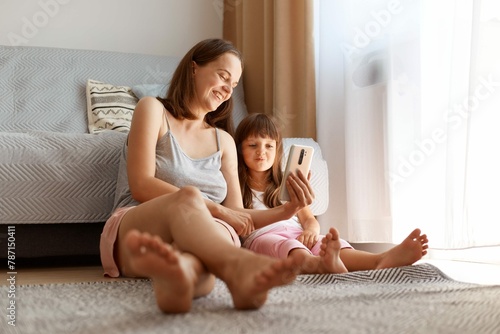 Positive Young Adult Caucasian Female Wearing Casual Style Clothing Posing Indoor Living Room With Her Daughter While Sitting Floor Near Sofa Window Watching Funny Video With Child