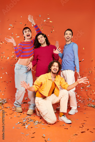 four positive stylish gay friends in bold outfits posing actively under confetti rain, pride month photo