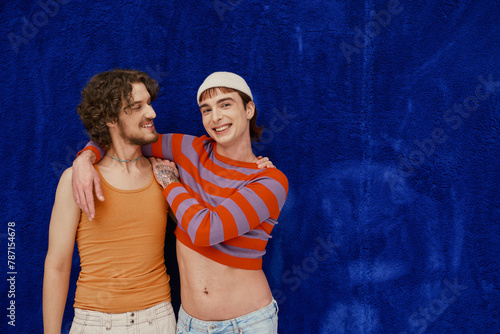 two jolly good looking gay men in vibrant clothes posing on dark blue backdrop, pride month © Bliss