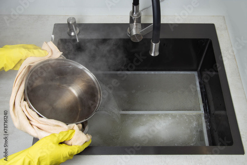 Man's hands in yellow gloves pour pots of hot water into a sink with dirty filters from the hood in the kitchen. Cleaning from dirt and grease with an oxygen cleaner. Copy space for text