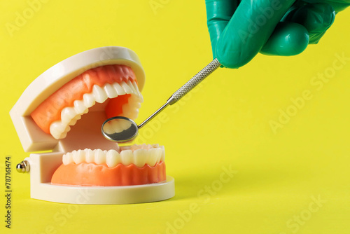 The hand of a dentist doctor in a green medical glove holds a dental mirror in his hand near a mock-up of a dental jaw on a yellow background. Concept of wisdom teeth removal, gum inflammation