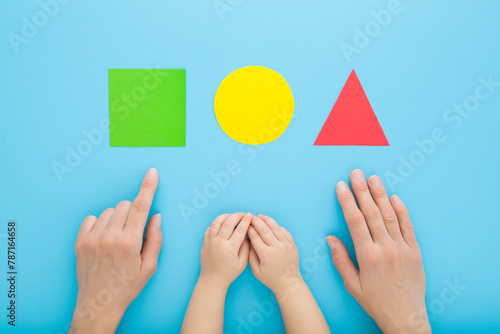 Young mother finger showing colorful geometric paper shapes to baby. Hands together on pastel blue table background. Time to learning. Infant development. Closeup. Point of view shot. Top down view.