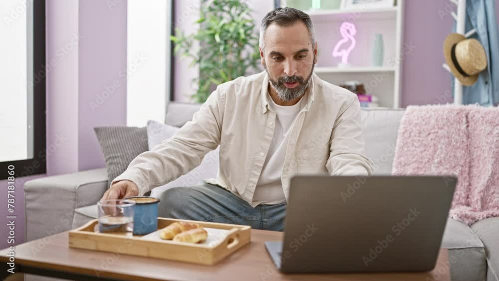 Mature bearded man using laptop in a cozy living room while enjoying a cup of coffee and breakfast. Mature bearded man using laptop in a cozy living room while enjoying a cup of coffee and breakfast.