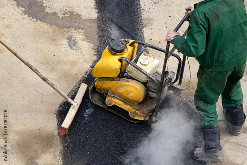 A road service worker compacts asphalt with a mechanical compactor.