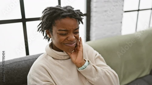 Young black woman wincing in tooth pain, nursing a dental ache while sitting at home on the sofa, hand to mouth, dreadlocks adding to the portrait of distress. photo