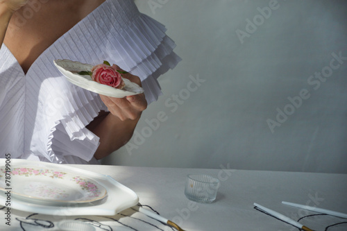 A woman, a girl in a stylish white dress holds a white porcelain saucer with a pink rose in her hands. Tea party, morning of the bride.