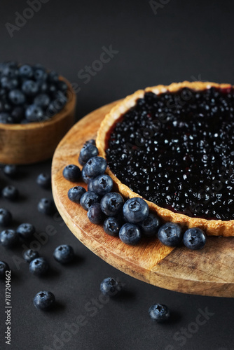 Opened blueberry tart on a wooden stand next to fresh blueberries on a dark background