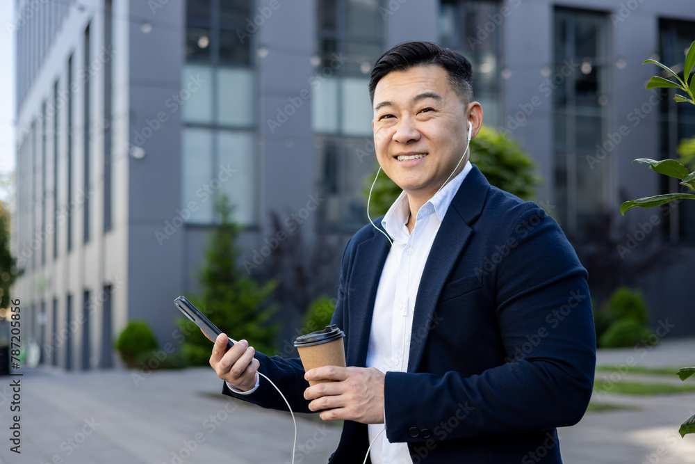 Portrait of an Asian young male office worker wearing headphones and holding a phone and a cup of coffee resting sitting outside an office center and smiling at the camera Portrait of an Asian young male office worker wearing headphones and holding a phone and a cup of coffee resting sitting outside an office center and smiling at the camera