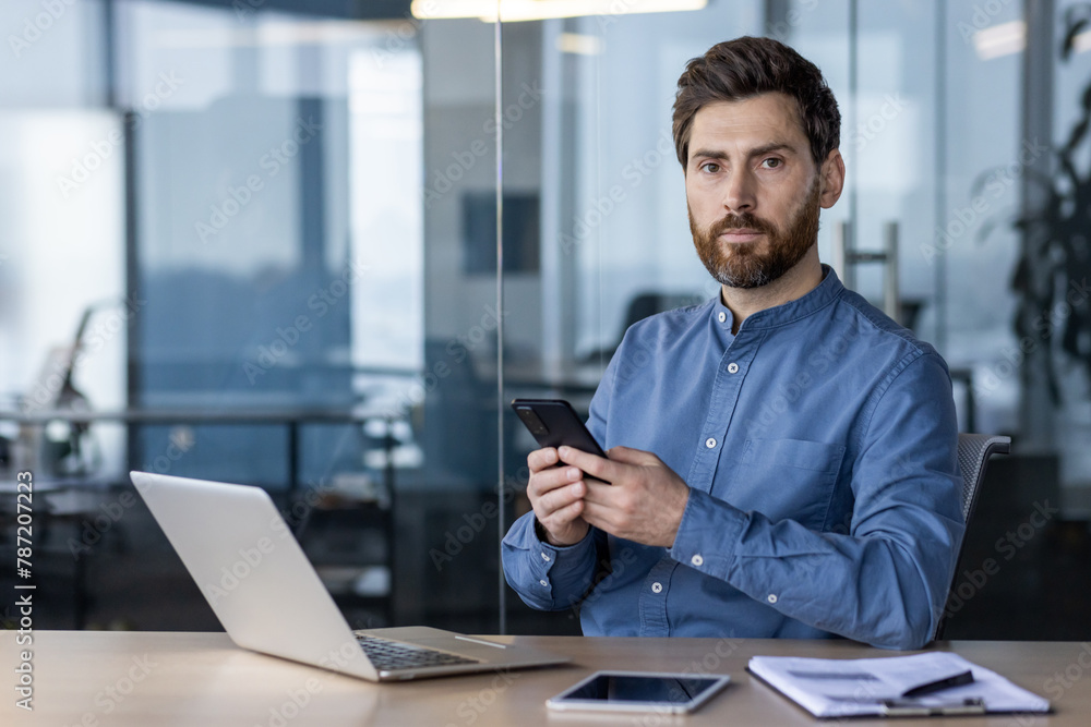 Portrait of a young serious man working in the office, sitting at a desk, holding a mobile phone and looking confidently into the camera Portrait of a young serious man working in the office, sitting at a desk, holding a mobile phone and looking confidently into the camera